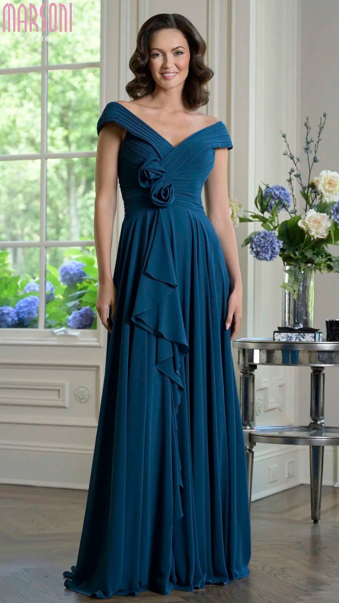 A woman stands in an elegant room wearing the Marsoni M333 long formal dress in teal, featuring off-the-shoulder chiffon with floral and ruffle details amid grand windows and beautiful floral arrangements.